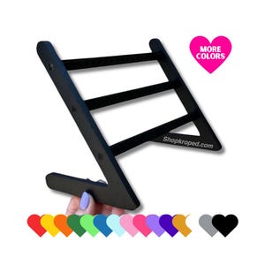 Peut inclure: Un support à boucles d'oreilles noir avec quatre rangées de chevilles pour ranger les boucles d'oreilles. Le support est incliné et a une base triangulaire. Le texte "Shopkropped.com" est imprimé sur le support. Un cœur rose avec le texte "PLUS DE COULEURS" est dans le coin supérieur droit de l'image.