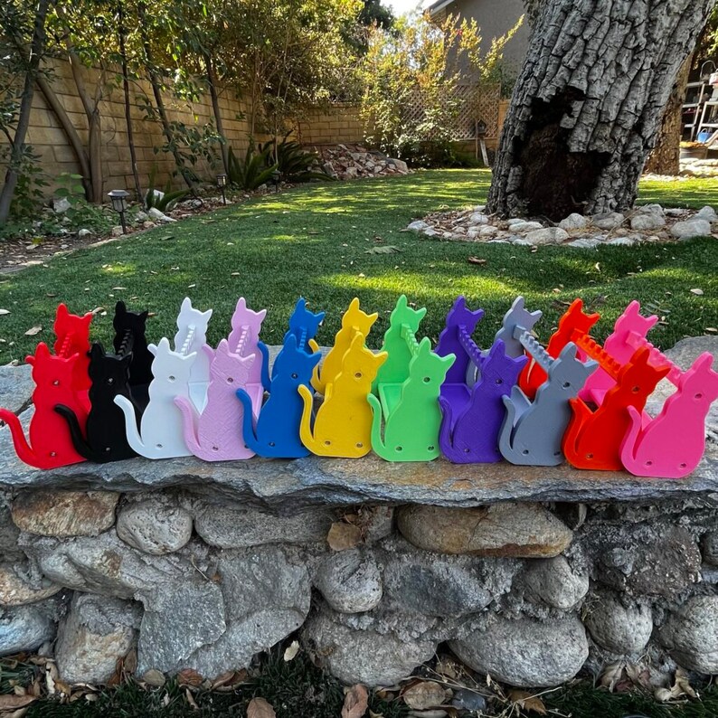 May include: A row of 12 colorful cat-shaped pencil holders, each with a different color. The colors are red, black, white, pink, blue, yellow, green, purple, gray, orange, and light blue.
