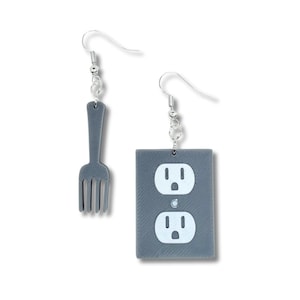 May include: A pair of quirky earrings. One earring is a gray fork, and the other is a gray rectangle with two white electrical outlets. Both earrings have silver-colored fish hook closures.