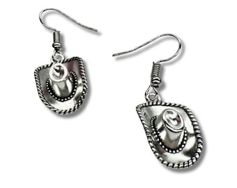 May include: A pair of silver-toned dangle earrings featuring a miniature cowboy hat charm. Each earring has a curved hook wire for attachment and a detailed hat design with a rope-textured brim and a central crown, finished in a metallic silver.
