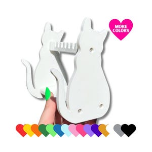 May include: White wooden wall mounted cat shaped holder with a comb for holding hair ties or other small items. The holder features two cats, one sitting on top of the other. The text "MORE COLORS" is in a pink heart shape.