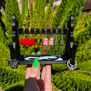 May include: A black jewelry stand with a variety of earrings, including red hearts, lightbulb shapes, gummy bear shapes, and skeleton shapes. There is also a black and white beaded necklace and two silver rings.
