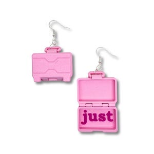 May include: Pink suitcase-shaped earrings with a silver hook. The suitcase is open and the inside says "just" in pink.