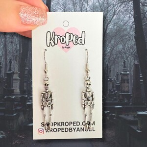 Puede incluir: Pendientes de esqueleto plateados colgados de una tarjeta blanca con el texto "Kroped by Angel". Los pendientes presentan un dise&ntilde;o de calavera y caja tor&aacute;cica. La tarjeta tambi&eacute;n muestra el sitio web shopkroped.com y el identificador de Instagram kropedbyangel.