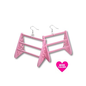 May include: A pair of pink earrings shaped like miniature A-frame structures. The earrings have a geometric design and are suspended from silver-colored ear wires. A pink heart with the text "MORE COLORS" is in the lower right corner.
