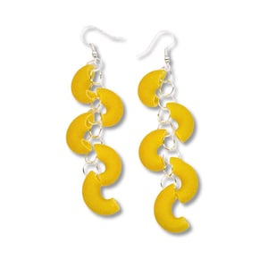 May include: A pair of unique dangle earrings featuring bright yellow macaroni noodles. Each earring has a silver-colored hook and chain, with four macaroni pieces dangling from the chain. The earrings are set against a white background.