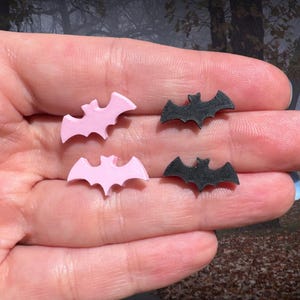 May include: Four small bat-shaped objects. Two are light pink, and two are black. The bats have a simple, stylized design with pointed ears and spread wings. The objects are likely decorative or craft supplies.