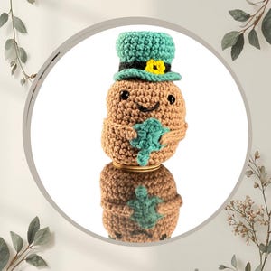 May include: A crocheted St. Patrick's Day themed can cozy in the shape of a leprechaun. The cozy is brown with a green hat and shamrock accents. The text "Cosy Cottage Co" and "@cosycottageco" are visible.