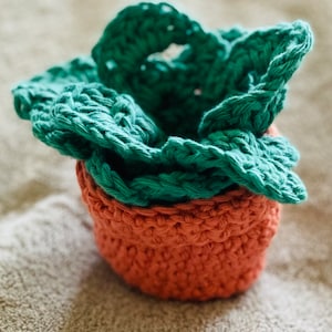 以下が含まれることがあります： 緑の葉っぱのデザインが施された、かぎ針編みで作った植木鉢。柔らかく、質感のある糸で作られており、自然で土っぽい雰囲気です。
