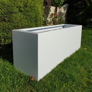 May include: A white rectangular planter box on green grass. The box has a simple, modern design and is made of a solid material. The planter is empty, ready for plants. The background features green bushes and a tree.