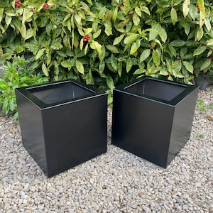 May include: Two black square planters made of metal. The planters are sitting on a bed of gravel.