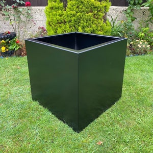 May include: A black square planter made of metal. The planter is sitting on a green lawn.