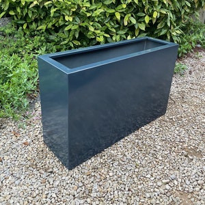 May include: A dark grey rectangular planter made of metal. The planter is sitting on a bed of small grey gravel.