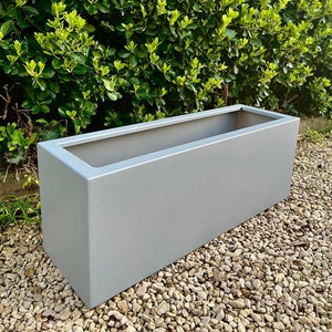 May include: A long, rectangular planter made of a light grey, textured material. The planter is sitting on a bed of small, grey stones.