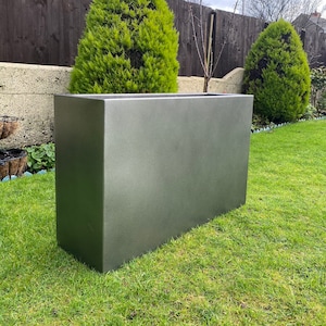 May include: A large, rectangular planter made of a dark grey, textured material. The planter is sitting on a green lawn in front of a wooden fence and a small evergreen tree.