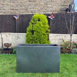 May include: A dark gray rectangular planter sits on a green lawn in front of a stone wall and a wooden fence. A small evergreen tree is growing inside the planter.