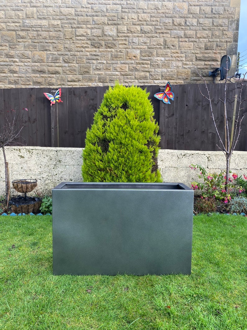 May include: A dark gray rectangular planter sits on a green lawn in front of a stone wall and a wooden fence. A small evergreen tree is growing inside the planter.