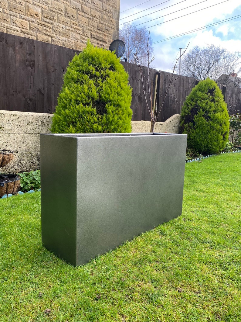 May include: A large, rectangular planter made of a dark gray, textured material. The planter is sitting on a green lawn in front of a wooden fence and a small evergreen tree.
