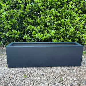 May include: A dark grey rectangular planter box. The box has a simple, modern design and is made of a solid material. It sits on a bed of small, light-coloured stones, with a backdrop of lush green foliage.