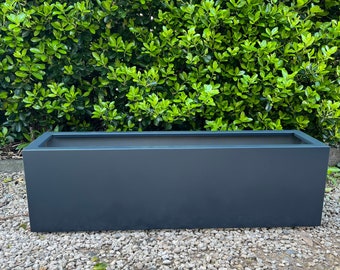 May include: A rectangular, dark gray planter box. The box is made of a solid material and has a simple, modern design. The planter is set on a bed of small, light-colored stones, with a backdrop of lush green foliage.