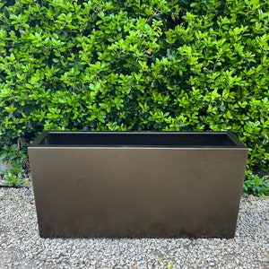 May include: A rectangular, dark brown planter made of metal. The planter is sitting on a bed of small, grey gravel.