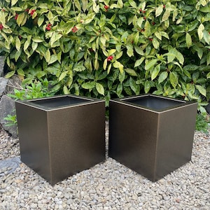 May include: Two bronze-coloured, square planters. The planters have a textured finish and are placed on a bed of small, light-coloured stones. The background features green foliage with red berries.