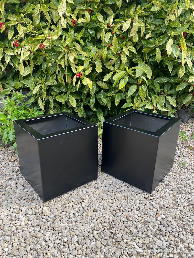 May include: Two black square planters made of metal. The planters are sitting on a bed of gravel.