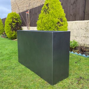 May include: A large, rectangular, dark gray planter made of concrete or a similar material. The planter is sitting on a green lawn in front of a house.
