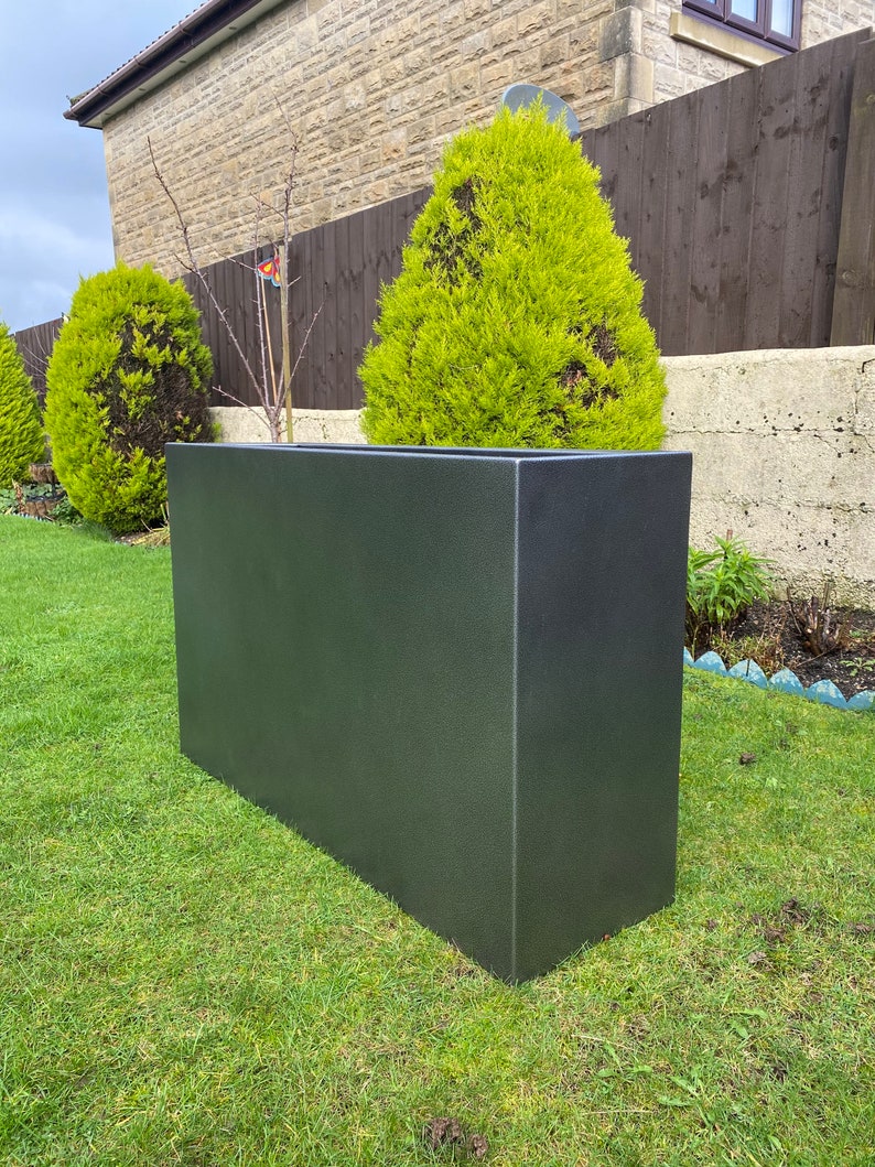 May include: A large, rectangular, dark gray planter made of concrete or a similar material. The planter is sitting on a green lawn in front of a house.
