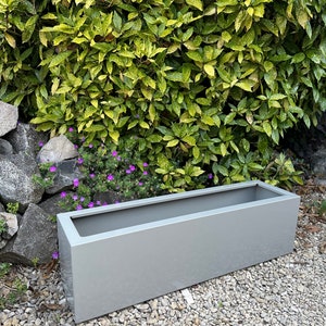 May include: A long, rectangular planter made of metal. The planter is a light grey colour and is sitting on a bed of gravel.