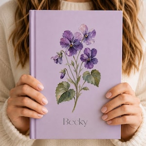 May include: A light purple hardback notebook featuring a watercolour illustration of purple pansy flowers with green leaves centred on the front cover. The name Becky is printed in a simple dark grey serif font below the floral artwork.