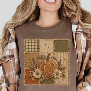 Könnte beinhalten: Ein espressofarbenes T-Shirt mit einem quadratischen Grafikdruck eines Kürbisses, der von Blumen und Blättern umgeben ist. Das Design besteht aus einem Raster im Patchwork-Stil mit verschiedenen Texturen, darunter karierte und gestreifte Muster.