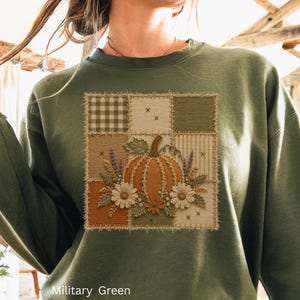 Può includere: Una felpa a maniche lunghe color verde militare con una grafica quadrata centrale dal design patchwork. L'illustrazione mostra una zucca arancione circondata da margherite bianche e rametti di lavanda su uno sfondo a quadri e righe.