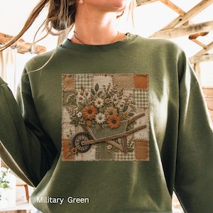 May include: A military green crewneck sweatshirt featuring a central square graphic of a wheelbarrow filled with orange and white flowers set against a patchwork background of tan, brown, and white checkered and solid fabric patterns.