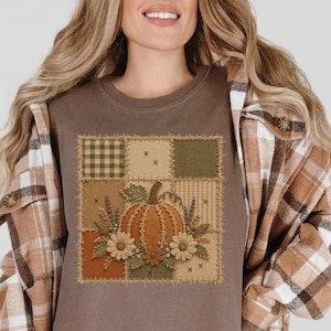 May include: An espresso-coloured t-shirt featuring a square graphic print of a pumpkin surrounded by flowers and foliage. The design is composed of a patchwork-style grid with various textures, including plaid and striped patterns in shades of brown and green.
