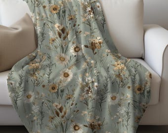 May include: A soft throw blanket featuring a sage green background with a repeating pattern of wildflowers, including white daisies, yellow blossoms, and brown dried grasses, arranged in a botanical illustration style across the fabric.