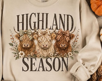 May include: A beige crewneck sweatshirt featuring a graphic of three Highland cows wearing patterned bows, surrounded by floral accents. The text HIGHLAND SEASON is printed in a dark brown serif font above and below the cows on the front chest.