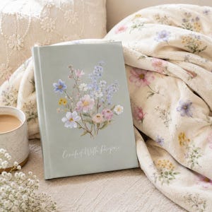 May include: A sage green hardcover journal featuring a delicate watercolour illustration of wildflowers in shades of blue, pink, and yellow. The text "Created With Purpose" is printed in white script at the bottom centre of the front cover.