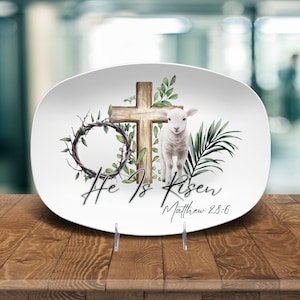 May include: White oval serving platter with a religious theme. The design includes a wooden cross, a lamb, a crown of thorns, and the text "He Is Risen Matthew 28:6" in script. Green foliage accents the design.