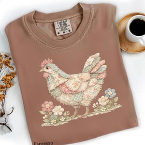 May include: Espresso-colored Comfort Colors t-shirt featuring a patchwork chicken design with floral accents. The shirt has a crew neck and short sleeves. A small tag is visible near the collar. A cup of coffee and flowers are in the background.