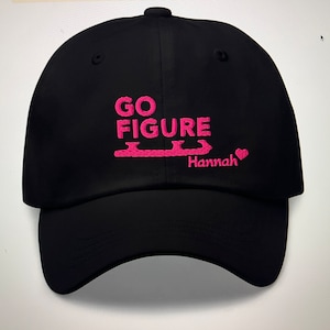 以下が含まれることがあります： ピンクの刺繍が施された黒いベースボールキャップ。キャップには「GO FIGURE」の文字と、スタイリッシュなスケートと「Hannah」という名前がピンクのハートと共に刺繍されています。
