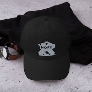 Puede incluir: Una gorra de béisbol negra con un gráfico bordado blanco de un jugador de hockey con la palabra "NOPE" en él.