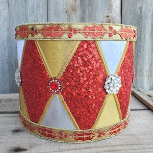 May include: A decorative drum-shaped container with a festive design. The container features a red sequined diamond pattern, gold and silver accents, and is trimmed with gold and red holly leaf detailing. Embellished with jewel-like accents.