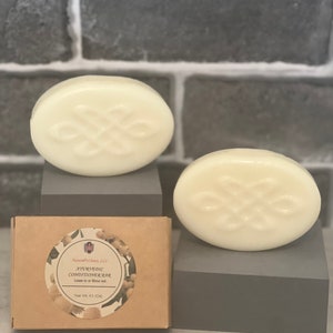 Pode incluir: Duas barras brancas de condicionador ayurvédico de forma oval, uma em cima de uma caixa marrom e a outra em cima de uma superfície cinza. A caixa está etiquetada com "Ayurvedic Conditioner" e "Leave-in or Rinse out". O peso líquido é de 4,5 oz.