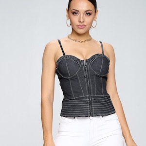 May include: A black denim corset top with a zipper closure and white stitching. The model is wearing white skinny jeans and black heeled sandals.