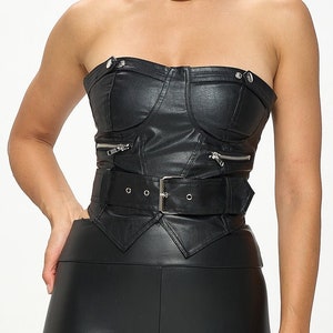 Puede incluir: Top negro sin tirantes de imitación de cuero con un corpiño ajustado, un cinturón de hebilla plateada y detalles de cremallera. El top tiene un dobladillo puntiagudo.