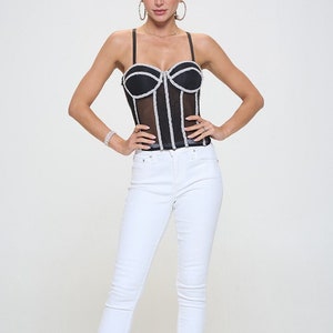 May include: A black mesh corset with white rhinestone trim and a sweetheart neckline. The corset is paired with white skinny jeans and sparkly silver heels.