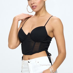 May include: A black mesh crop top with a sweetheart neckline and adjustable straps. The top has a fitted bodice and a loose, flowing bottom. The model is wearing white jeans.