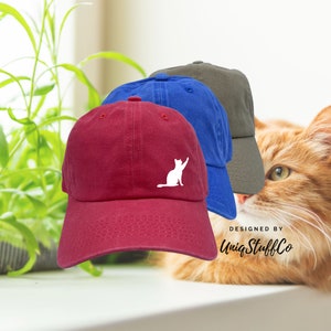 Puede incluir: Tres gorras de béisbol con una silueta blanca de un gato en la parte delantera. Las gorras son roja, azul y gris. El texto "DESIGNED BY UniqStuffCo" es visible en la esquina inferior derecha de la imagen.