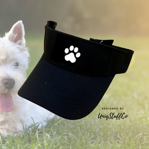 Op de afbeelding: Zwarte visor met een witte pootprint op de voorkant. De tekst "DESIGNED BY UniqStuffCo" staat rechtsonder op de visor.